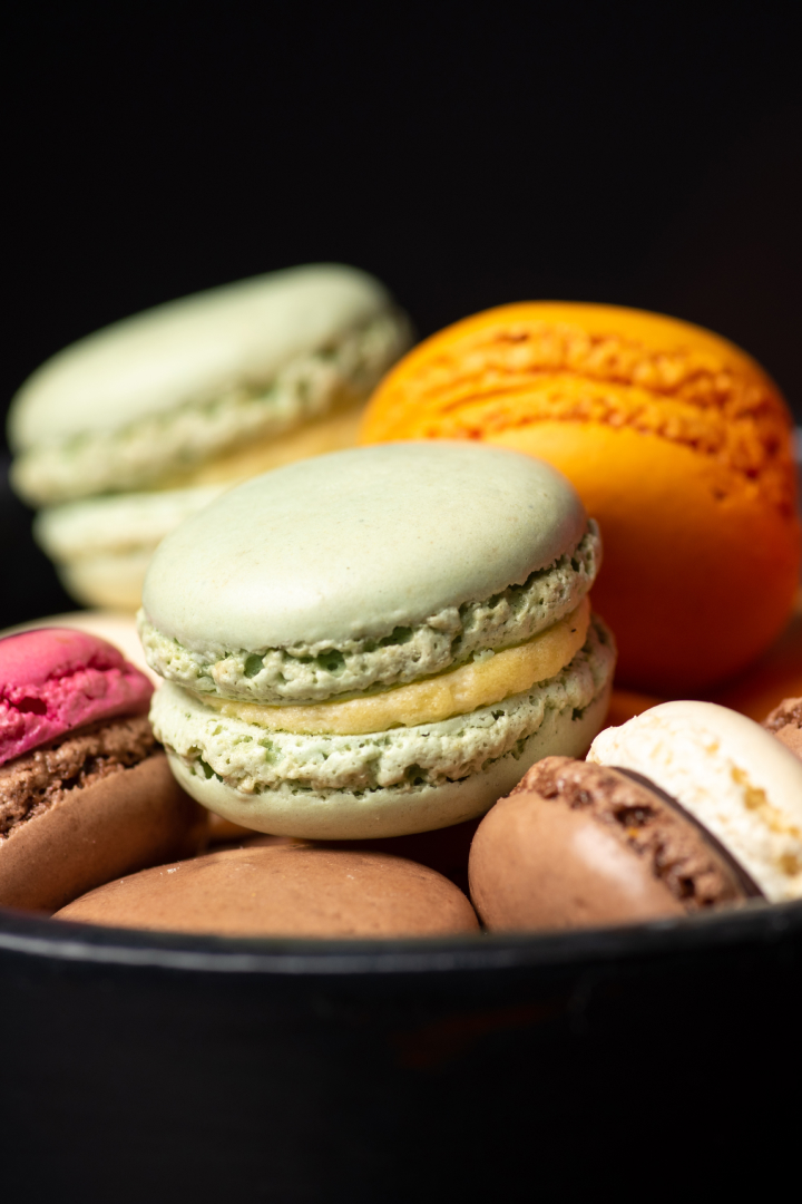 Des macarons colorés de différentes saveurs, comprenant du vert, du jaune, de l'orange, du rose et du marron, présentés sur un fond noir, mettant en valeur leur texture lisse et leur garniture crémeuse.