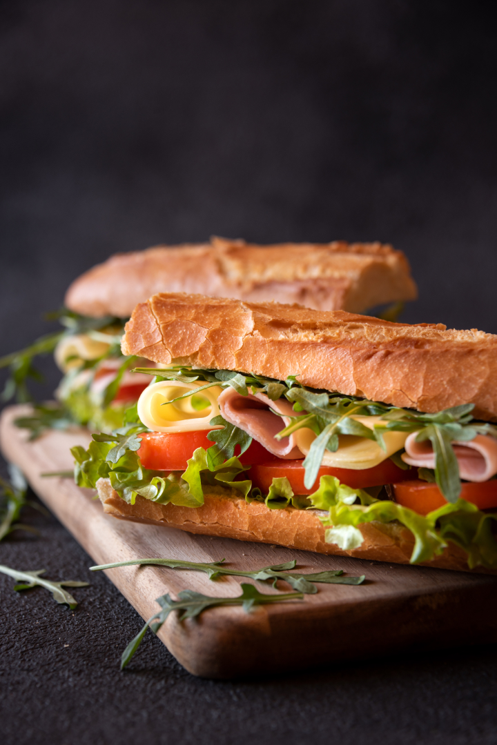 Un sandwich baguette garni de jambon, fromage, tomates, laitue et roquette, présenté sur une planche en bois avec un fond sombre.